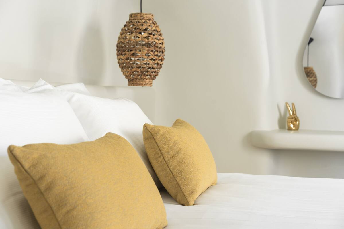 A bed with white linens and two mustard-colored pillows. A woven pendant light hangs above the bed.
