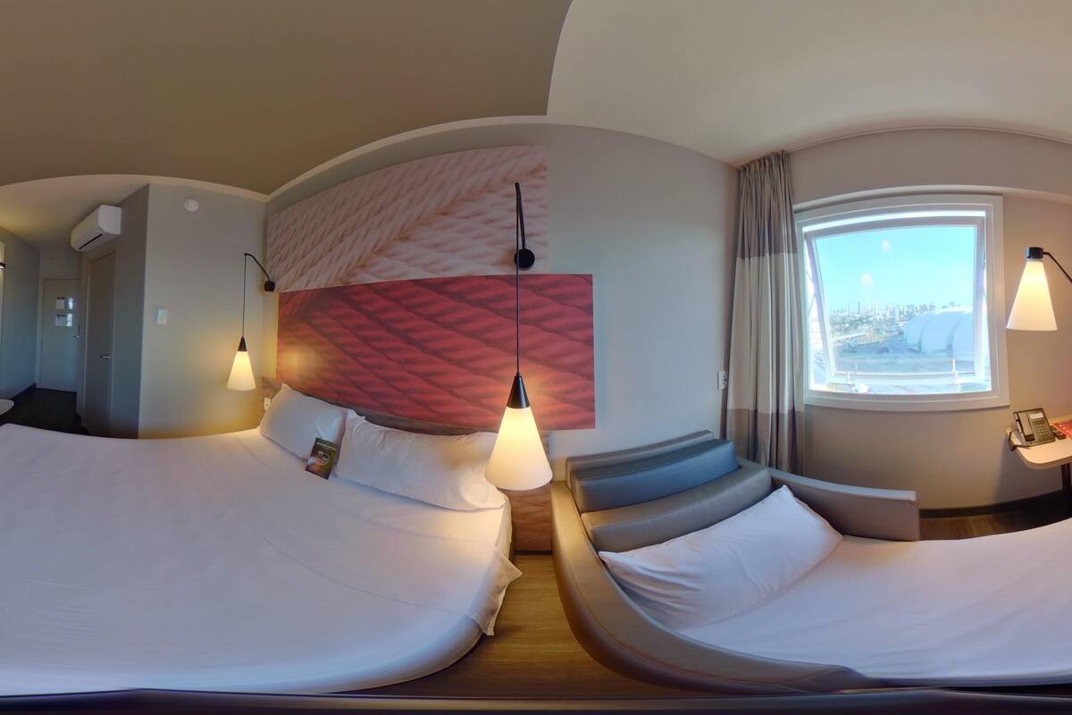 Une chambre d’hôtel avec deux lits, une grande fenêtre donnant sur la ville, un climatiseur fixé au mur et une lampe de chevet.