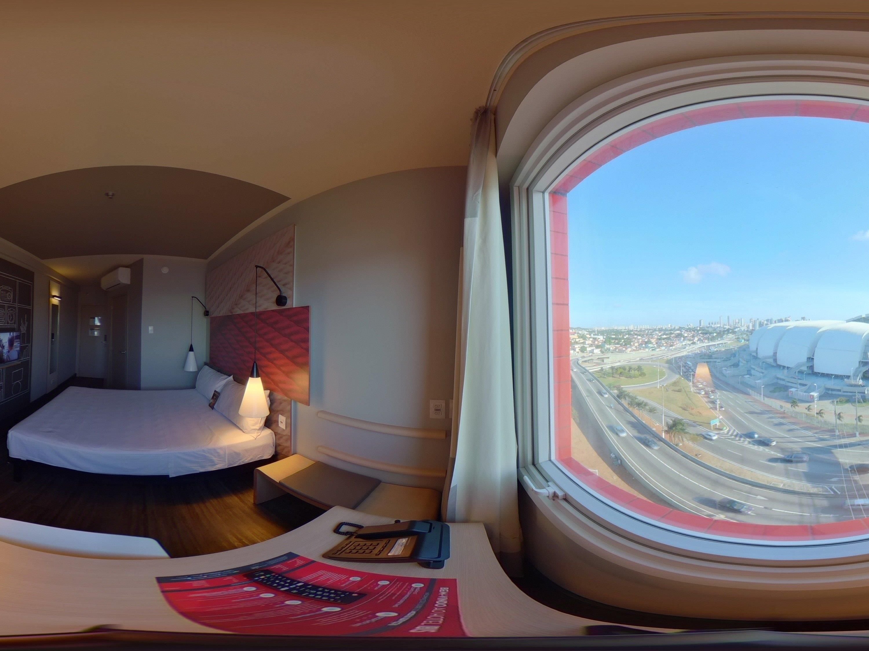 Quarto de hotel com cama, mesa de cabeceira, abajur e vista para a cidade através de uma janela grande.