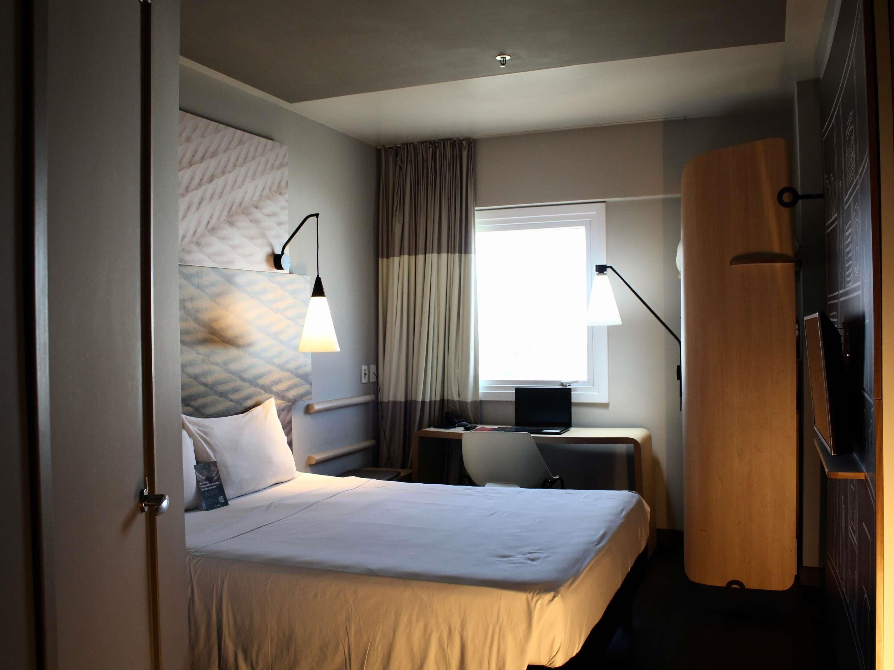 A hotel room with a bed, a desk, a laptop, a window with curtains, and a wooden wardrobe.