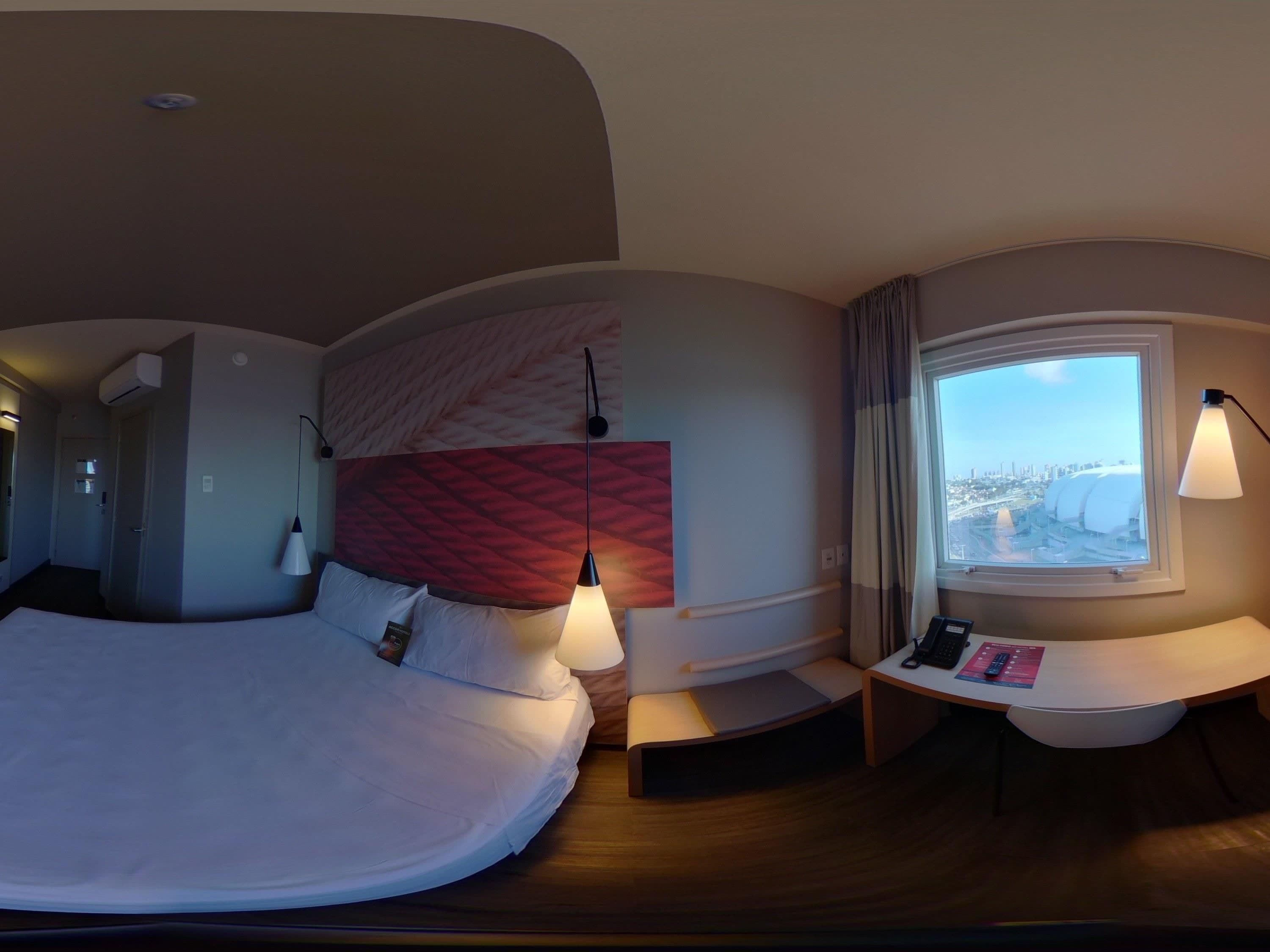 A modern hotel room with a curved bed, a headboard with a textured pattern, a bedside table with a lamp, a small desk with a remote control, and a window with a city view.