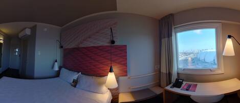 A modern hotel room with a curved bed, a headboard with a textured pattern, a bedside table with a lamp, a small desk with a remote control, and a window with a city view.