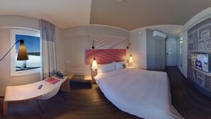 A modern hotel room with a curved desk, a bed with a white bedspread, a headboard with a pink pattern, and a view of the sky through a window.