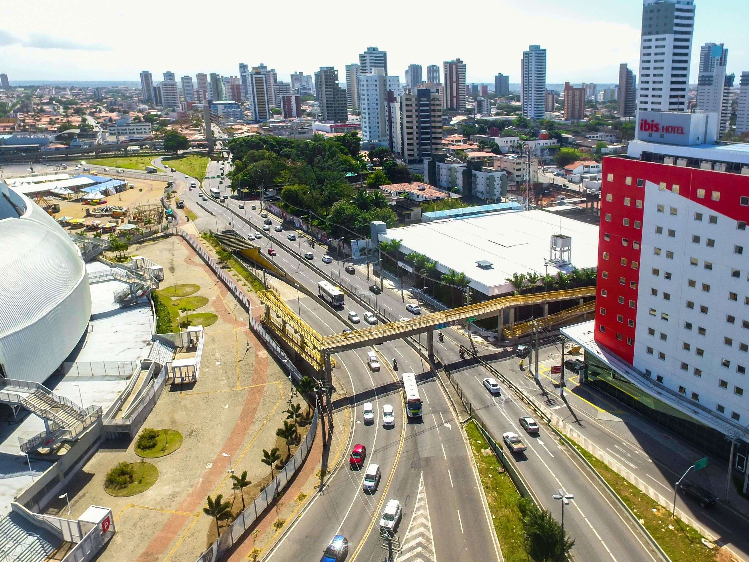 Paisagem urbana com um edifício vermelho e branco de destaque, uma grande estrutura em forma de cúpula branca e uma movimentada via com várias faixas e uma passarela para pedestres.