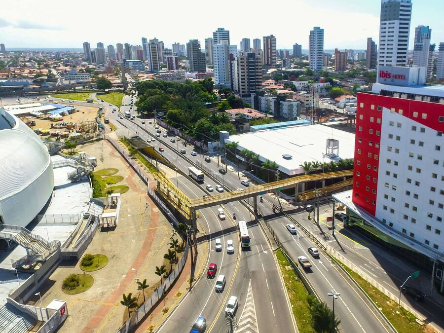 Paisagem urbana com um edifício vermelho e branco de destaque, uma grande estrutura em forma de cúpula branca e uma movimentada via com várias faixas e uma passarela para pedestres.