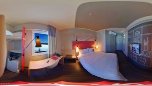 Habitación de hotel con cama, escritorio, silla, lámpara, televisor y minibar.