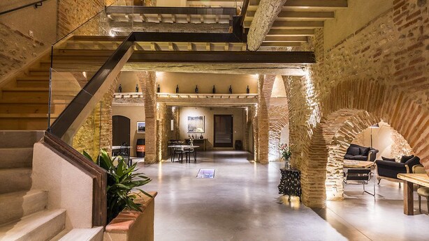 Un interior espacioso con una escalera central, paredes de piedra y una zona de estar con muebles modernos.