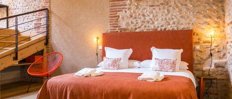 Un dormitorio con pared de ladrillo, escalera de madera, una silla roja y una cama con almohadas blancas y estampadas.