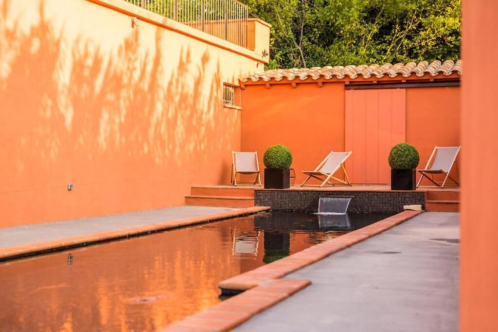 A swimming pool with two lounge chairs and potted plants, surrounded by orange walls and a tiled roof.