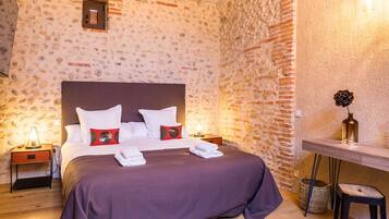 Un dormitorio con pared de piedra, cabecera de madera, mesita de noche con lámpara y escritorio con un florero.