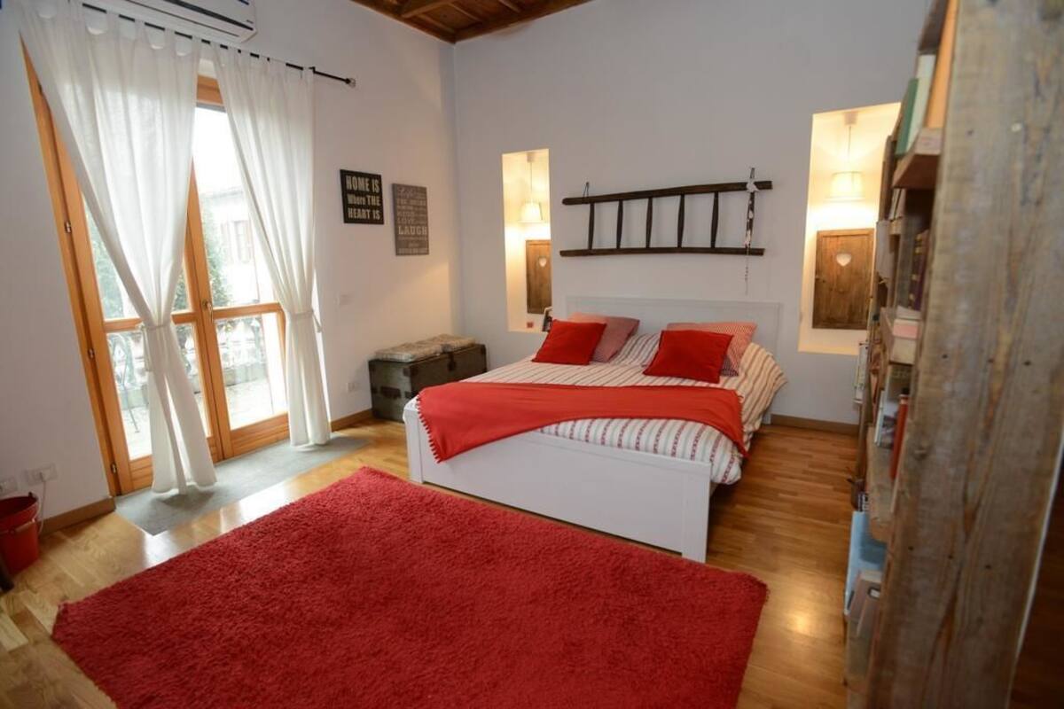 Ein Schlafzimmer mit einem Bett, roter Bettwäsche, Holzboden, einem roten Teppich, einem Fenster mit Vorhängen und einem an der Wand befestigten Regal.