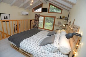 A bedroom with a bed on a wooden platform, a window with a view of greenery, a mounted TV, and a bedside lamp. - Il Pettirosso (Certosa di Pavia)