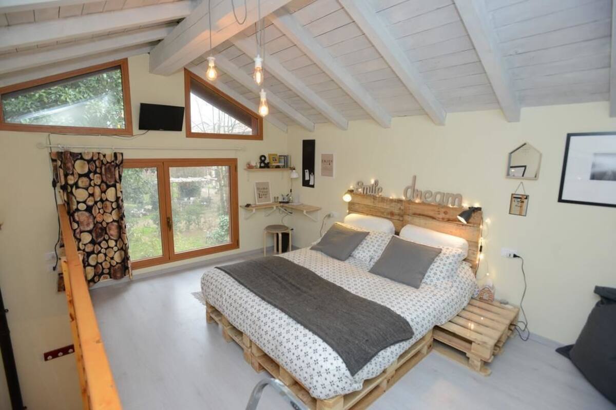 Ein Schlafzimmer mit einem Bett auf einer Holzlade, einem Fernseher an der Wand und einem Fenster mit Vorhängen.