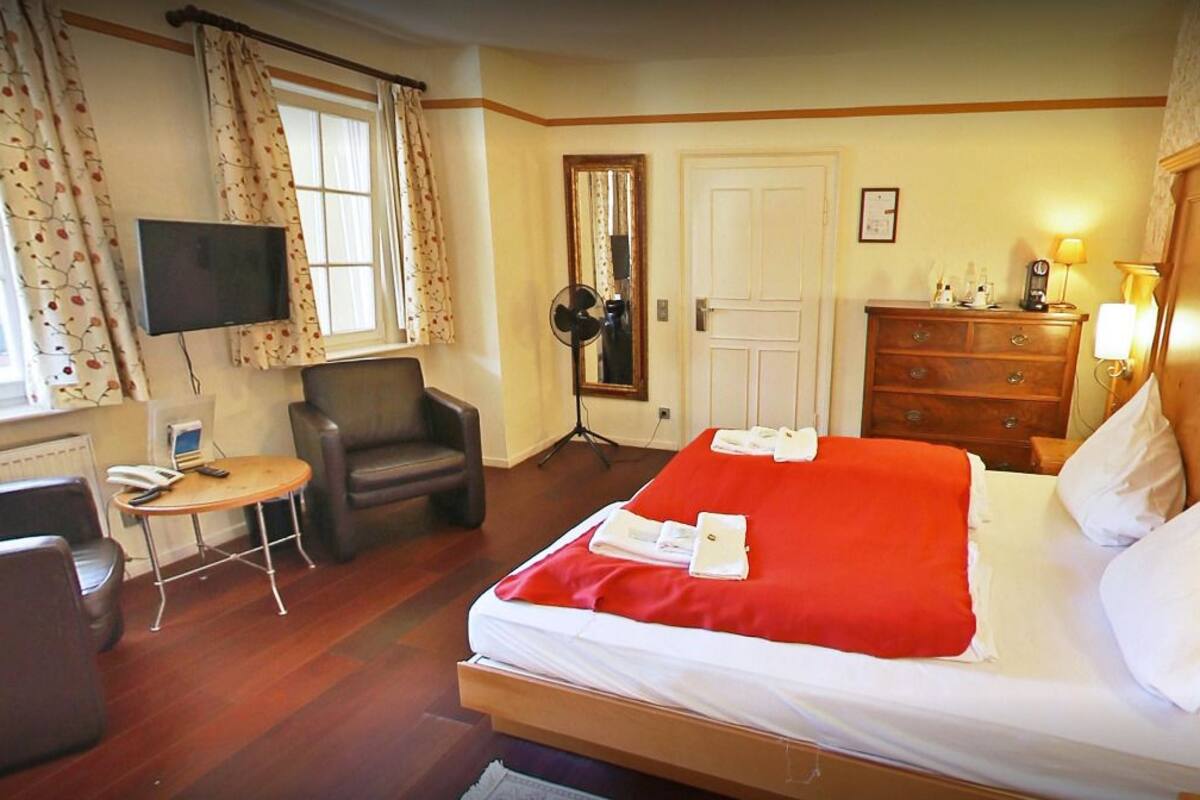 A hotel room with a bed, a desk, a chair, a TV, a dresser, and a fan.