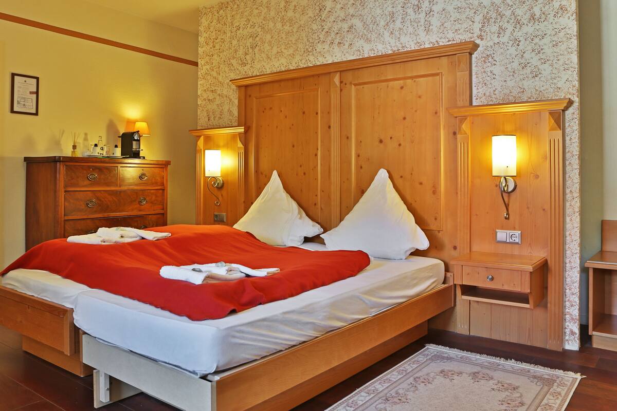 A neatly made bed with a red blanket and white pillows, wooden headboard, bedside tables with lamps, and a wooden dresser.