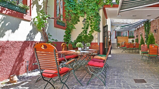 Een terras met houten tafels en stoelen, omgeven door een geplaveid pad en groen.