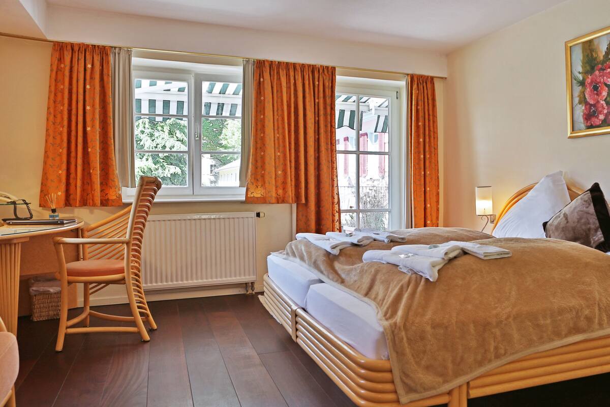 A hotel room with a wooden bed, a desk with a chair, a radiator, and a window with curtains.