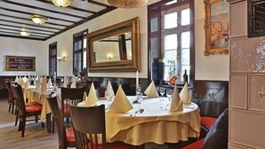 Een restaurant met ronde tafels, gedekt met witte servetten, kaarsen en wijnglazen. Er zijn houten stoelen en een grote spiegel aan de muur.