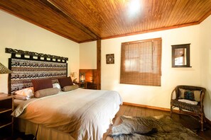 Ein Schlafzimmer mit einem großen Bett, Holzboden, einem Kopfende mit einem gemusterten Design, einem Fenster mit Jalousien, einem Sessel und einem Teppich.