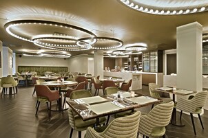 Um restaurante moderno com mesas redondas, cadeiras estofadas verdes e luminárias circulares no teto.