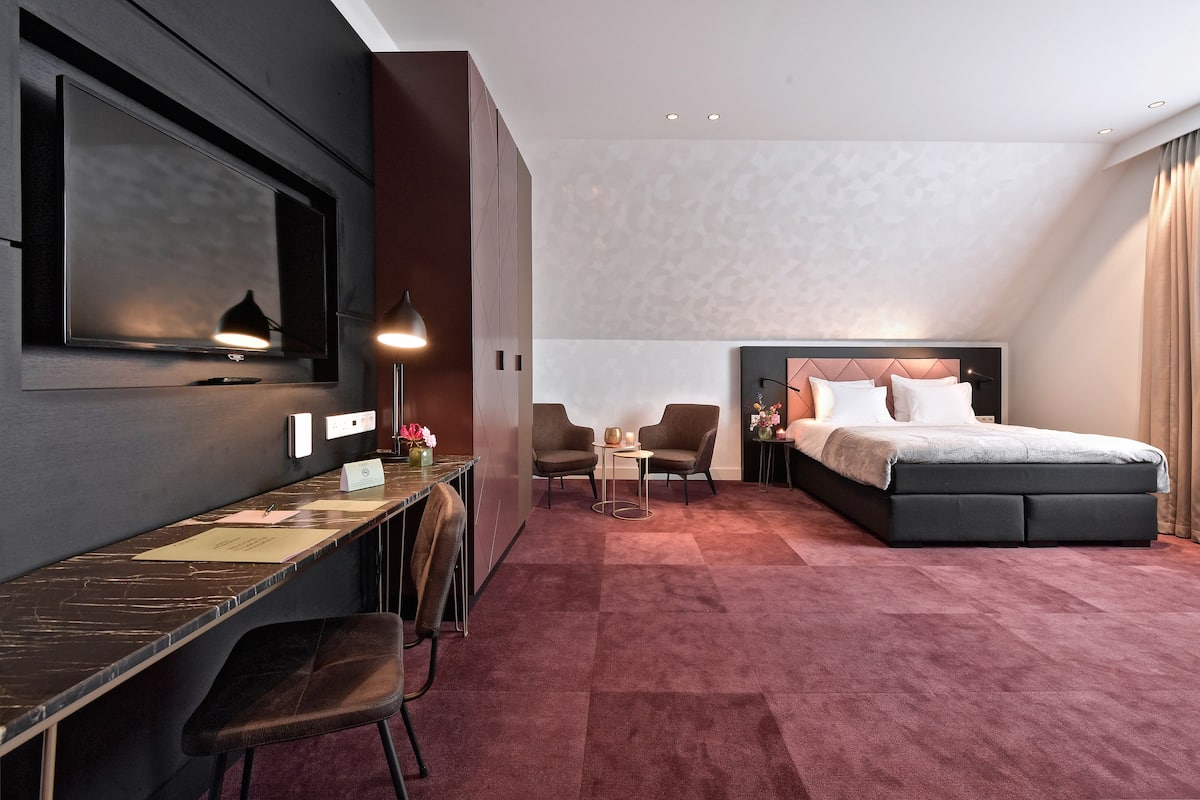 Una habitación de hotel moderna con una cama grande, un escritorio con una silla, un televisor montado en la pared y una pequeña mesa con dos sillas.