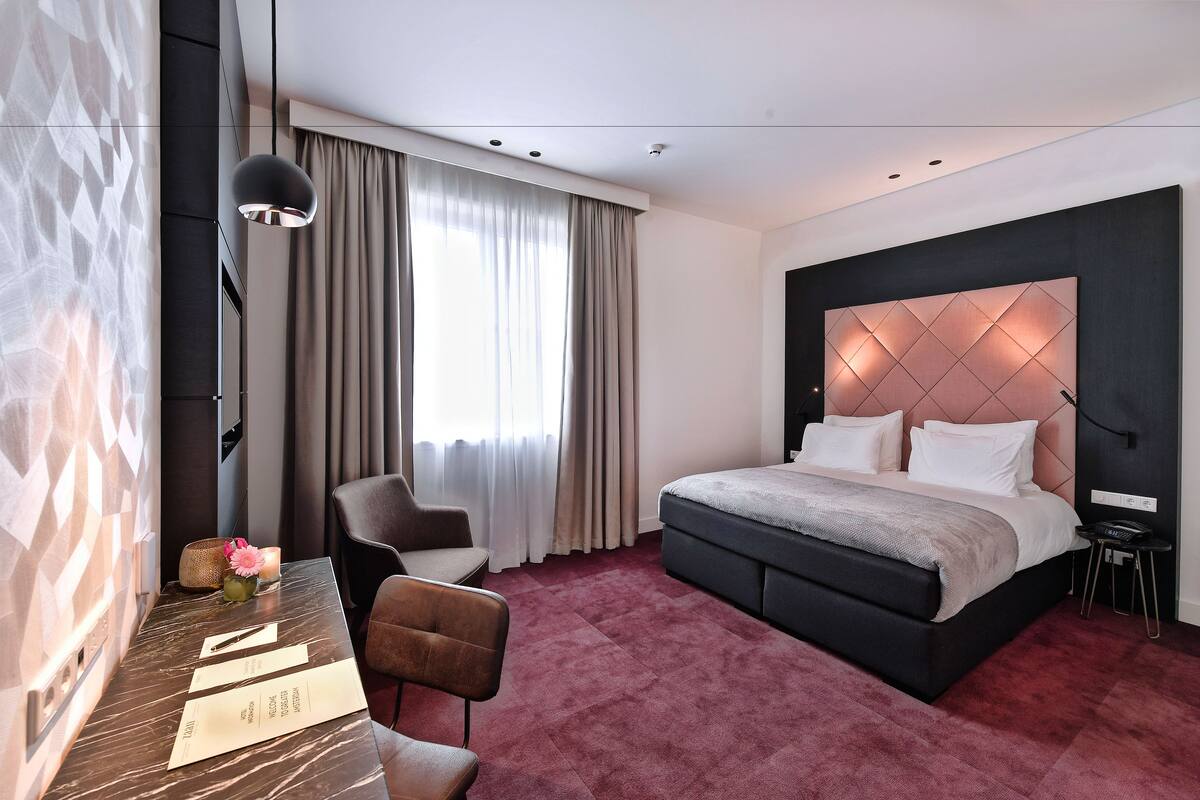 Una habitación de hotel moderna con una cama grande, un escritorio con una silla, una mesita con un florero y una vela, y un ventanal con cortinas.
