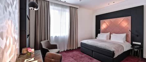 Habitación de hotel moderna con una cama grande, un escritorio con silla, una mesita con un florero y una vela, y una ventana con cortinas.