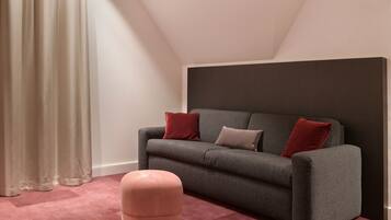 A modern living room with a grey sofa, two red cushions, a pink ottoman, and a large dark headboard.