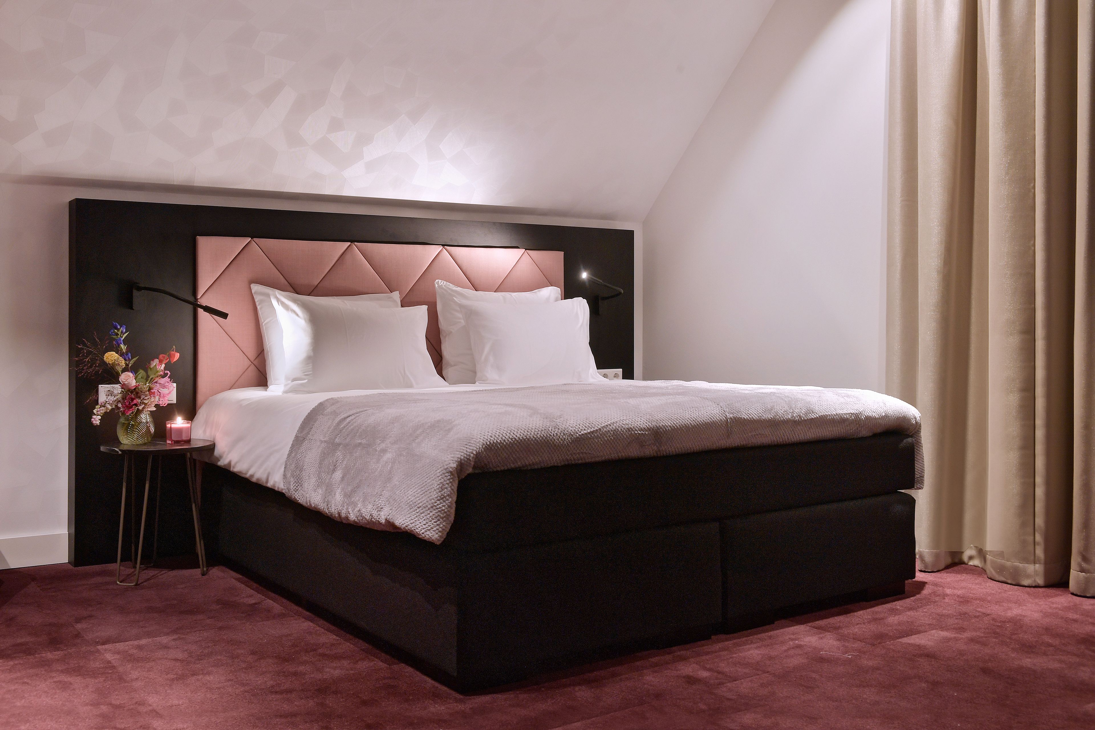 A modern bedroom with a dark headboard, white bedding, and a small bedside table with a vase and candle.