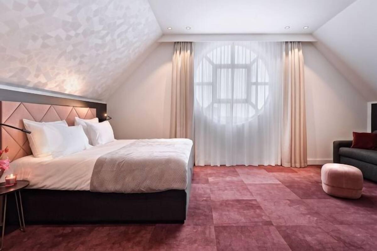 Un dormitorio con cama, mesita de noche, silla y un amplio ventanal con cortinas.