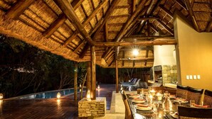 An outdoor dining area with a thatched roof, stone pillars, and a pool in the background. - The Island River Lodge (Hoedspruit)