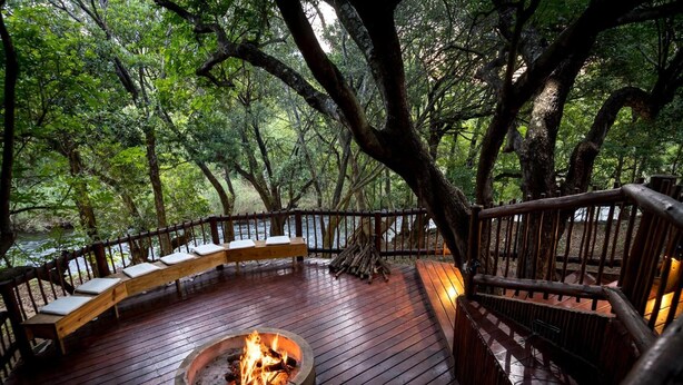 A wooden deck with a fire pit, benches, and a railing overlooking a forested area. - The Island River Lodge (Hoedspruit)