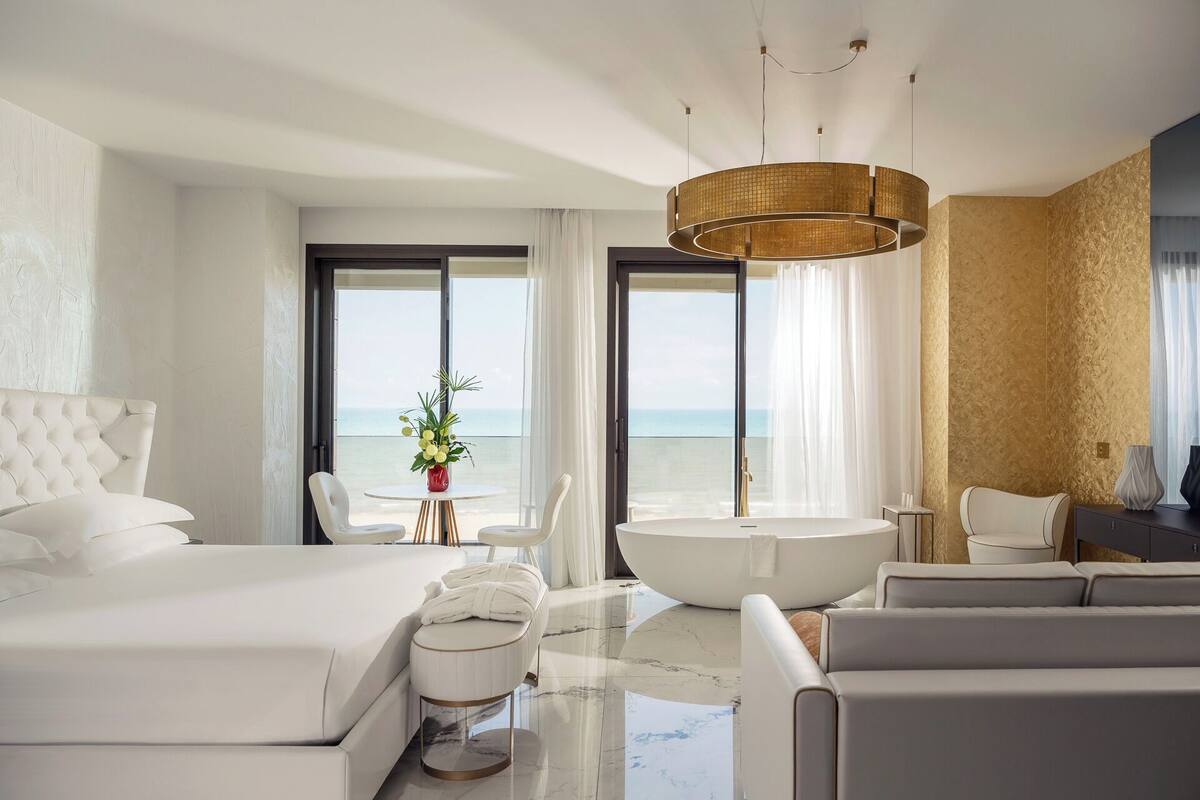 A modern hotel room with a large bed, a seating area with a view of the sea, a bathtub, and a large window.
