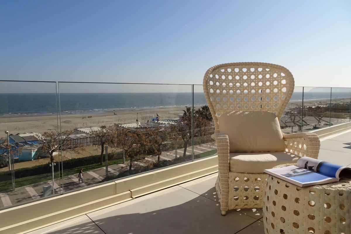 A balcony with a wicker chair and a small table, overlooking a beach and cityscape.