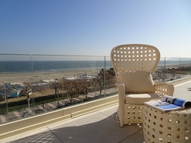 Un balcón con una silla de mimbre y una mesita pequeña, con vistas a la playa y al paisaje urbano.