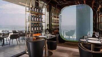 Un restaurante moderno con vista panorámica al océano, que cuenta con sillas de cuero negro, una barra de vinos con estantes llenos de botellas y un separador de vidrio.