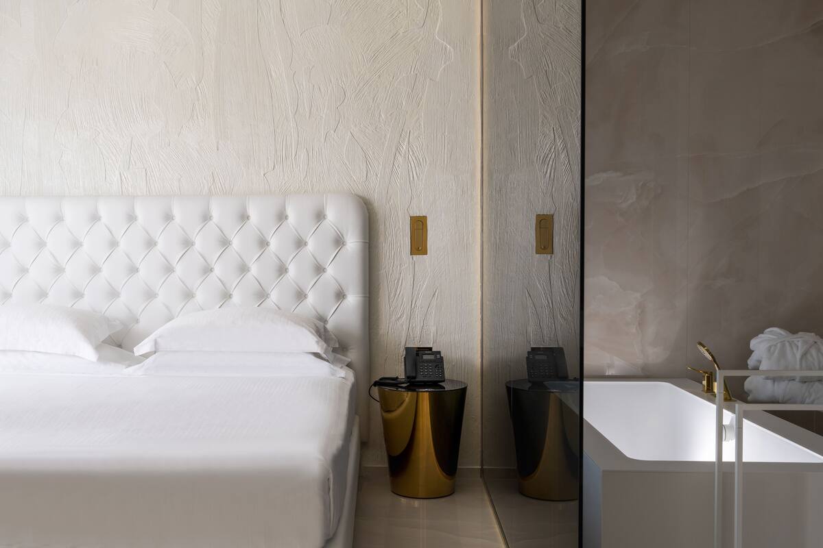 A modern hotel room with a tufted headboard bed, a gold side table, a black telephone, and a white bathroom with a large mirror and a gold faucet.