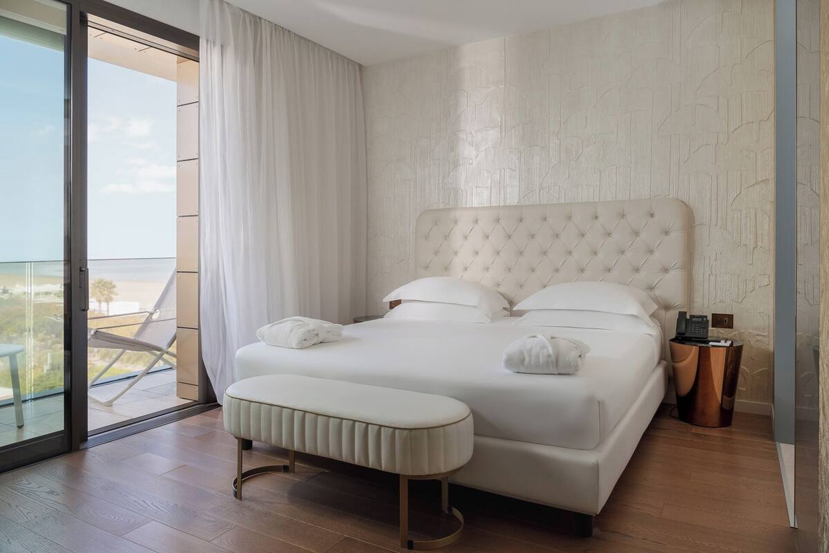 A bedroom with a large bed, a bench, a bedside table, and a view of the outdoors.