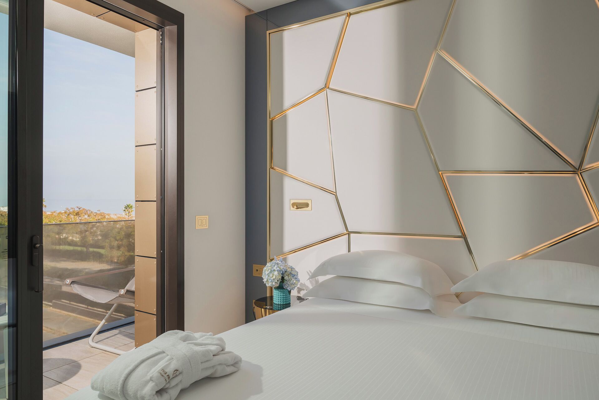 A modern hotel room with a geometric headboard, a bed with white linens, a small side table with a vase of flowers, and a view of the outdoors through a glass door.