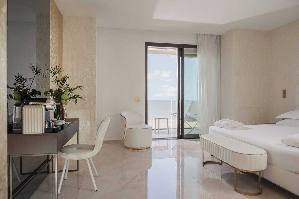 A modern hotel room with a bed, a desk, a chair, a small table, and a balcony with a view.