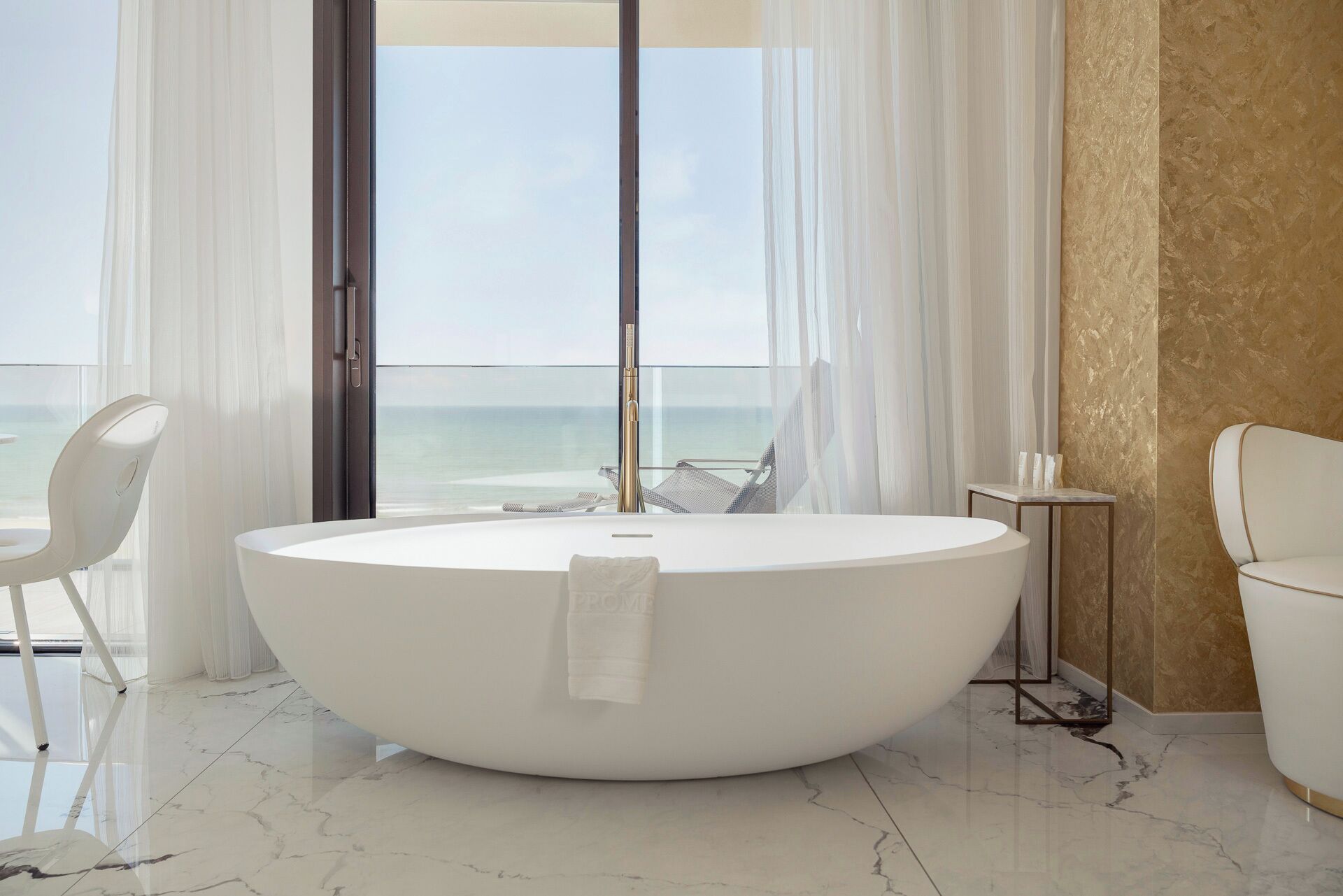 A modern bathroom with a large oval bathtub, a white chair, a small table, and a view of the ocean through large windows.