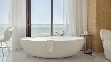 Une salle de bain moderne avec une grande baignoire ovale, une chaise blanche, une petite table et une vue sur l’océan grâce à de grandes fenêtres.