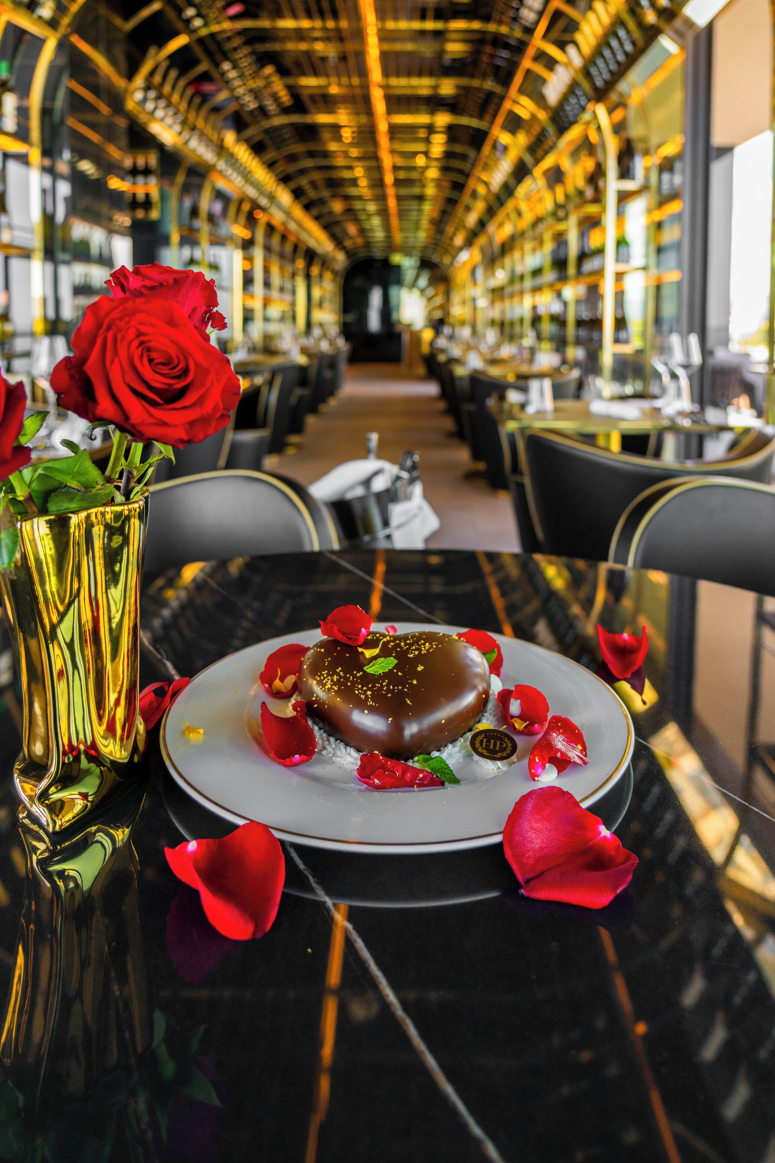 Una mesa de comedor con un postre, un jarrón con rosas rojas y una superficie de mesa negra reflectante.