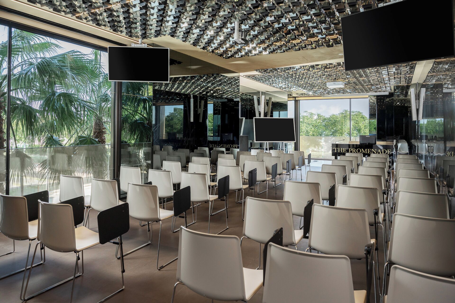 A conference room with rows of chairs, a large screen, and a glass wall with a view of palm trees.