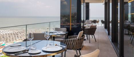 A terrace with round tables set for dining, overlooking a beach and the sea.
