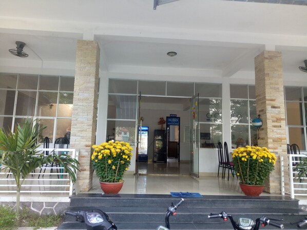 A hotel entrance with a glass door, two potted yellow flowers, and a motorbike parked in front. - Quang Nam University Guesthouse (Da Nang)