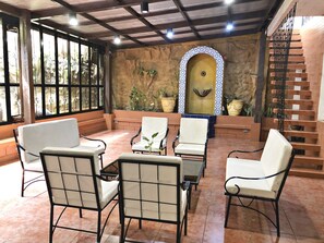Un patio iluminado por el sol con sillas blancas acolchadas, una mesa de vidrio y una fuente decorativa.