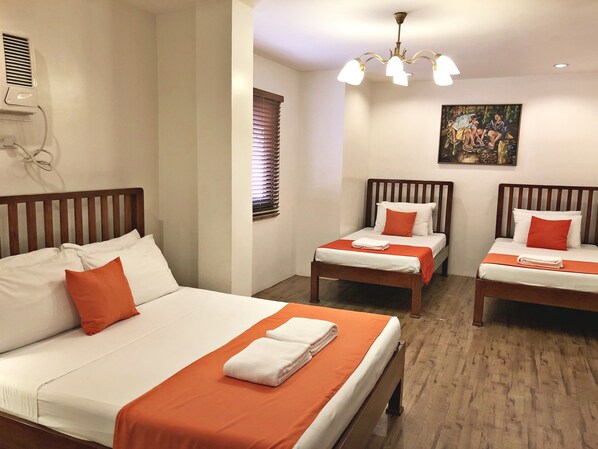 Habitación de hotel con dos camas individuales y una cama doble, cada una con ropa blanca y detalles naranjas. Hay un cuadro en la pared, una lámpara de techo y una ventana con persianas.