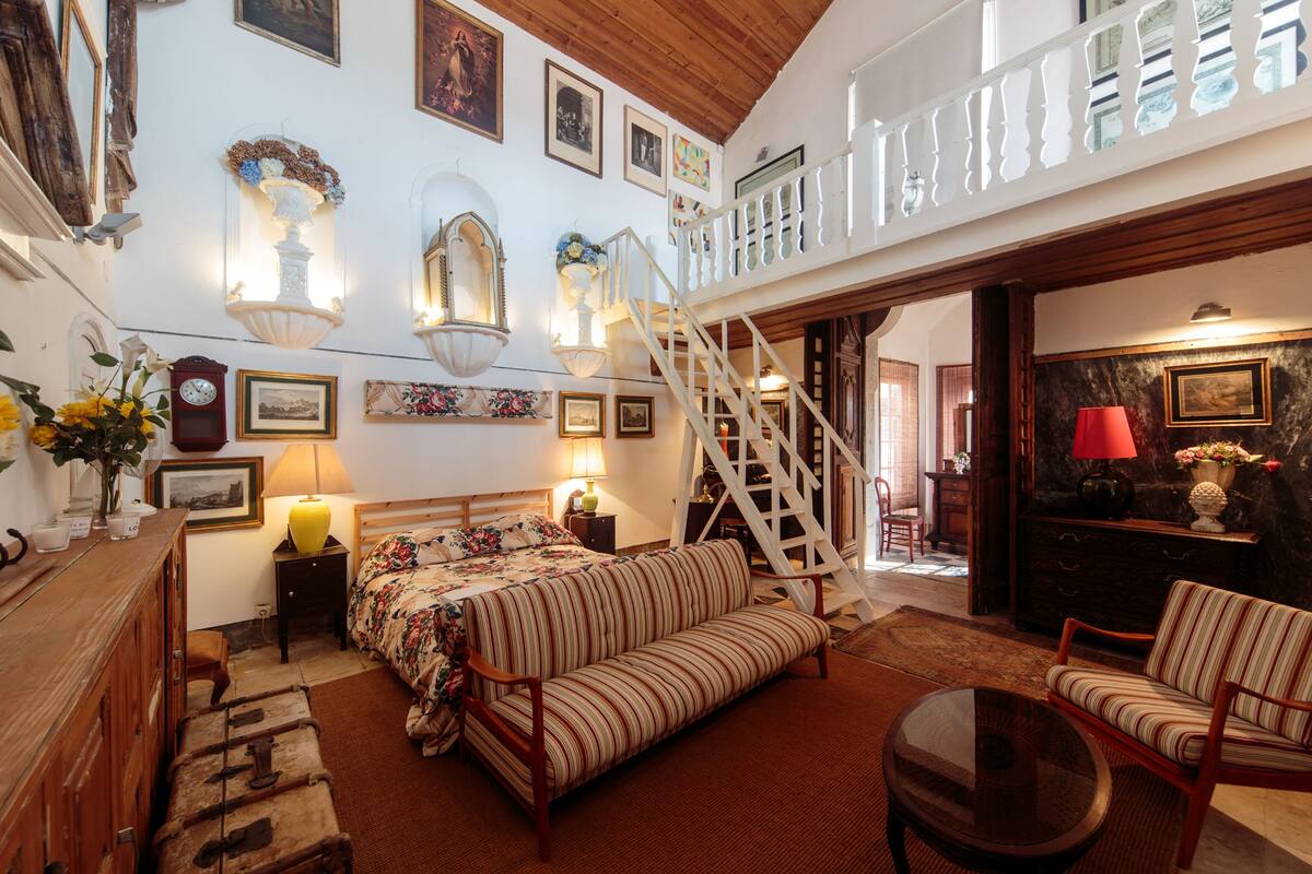 A spacious room with a large bed, a sofa, a dining table, and a staircase leading to an upper level.
