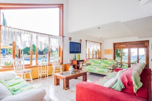 A spacious living room with a large window, a red sofa, a wooden coffee table, and a television mounted on the wall.